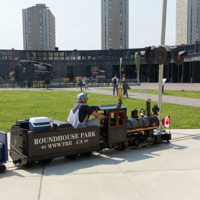 Der Lokführer kreist mit seiner geschrumpften Dampflok vor einer ehemaligen Remise in Toronto, mitfahren möglich.